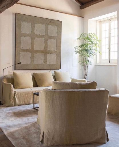 May include: A living room with a beige sofa and two matching armchairs. The sofa has three pillows and a coffee table in front of it. A large abstract painting hangs on the wall behind the sofa. A potted plant is in the corner of the room.