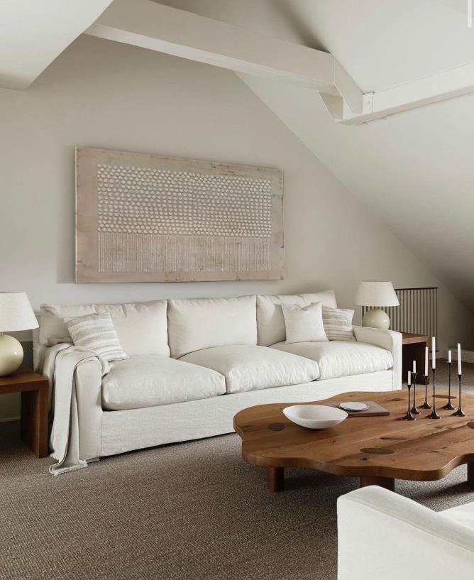 May include: A large rectangular wall art piece with a textured pattern of white dots and vertical lines, positioned above a cream-colored three-cushion sofa. The sofa features a draped throw blanket, two side tables with lamps, and a wooden coffee table.