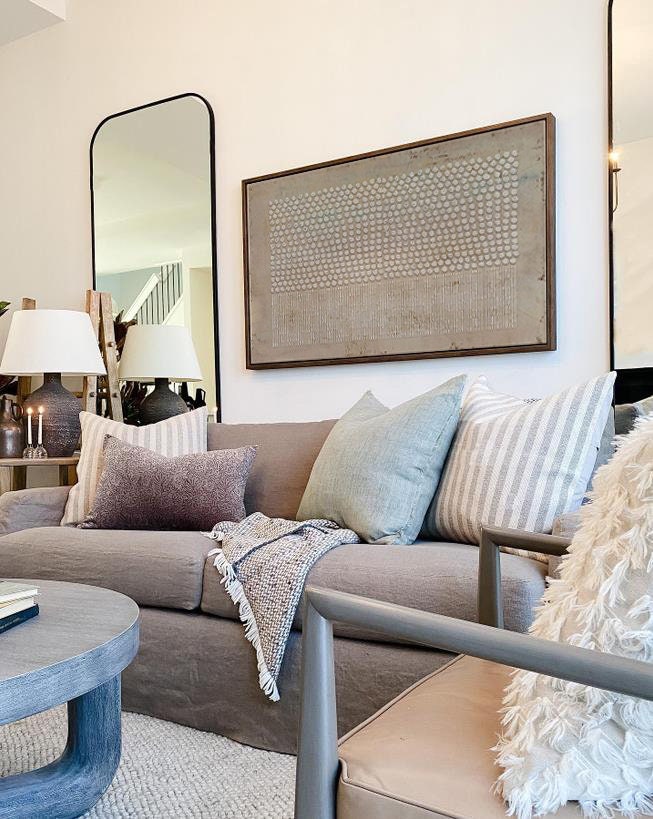 May include: A gray sofa featuring a collection of throw pillows in solid sage green, white and gray stripes, and a textured dark purple. A patterned throw blanket with fringe rests on the seat. Above the sofa hangs a large rectangular wall art piece.