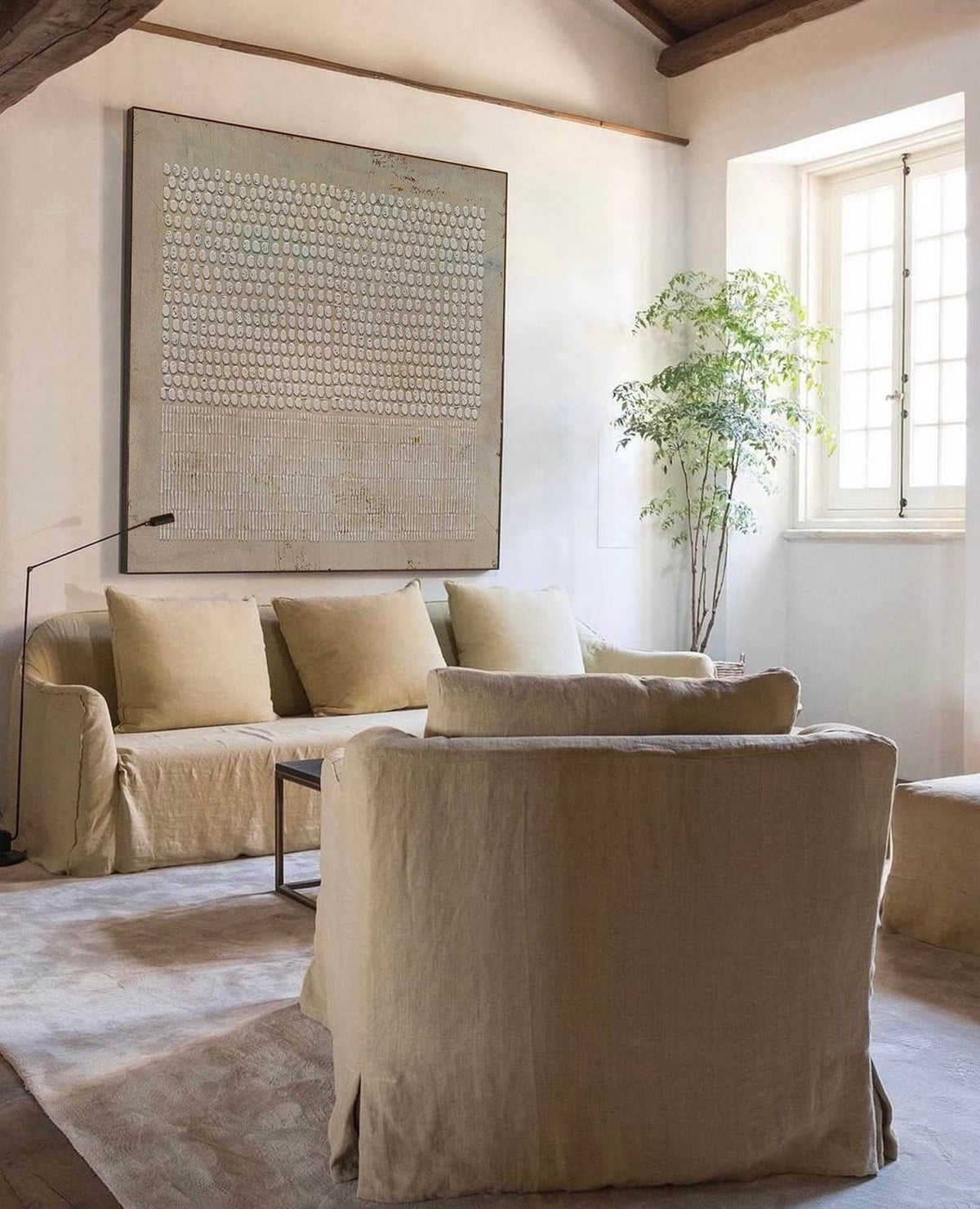 May include: A living room with a light beige sofa and two matching armchairs. The sofa has three pillows and a small coffee table in front of it. A large abstract painting hangs on the wall behind the sofa. A potted plant is in the corner of the room next to a window.