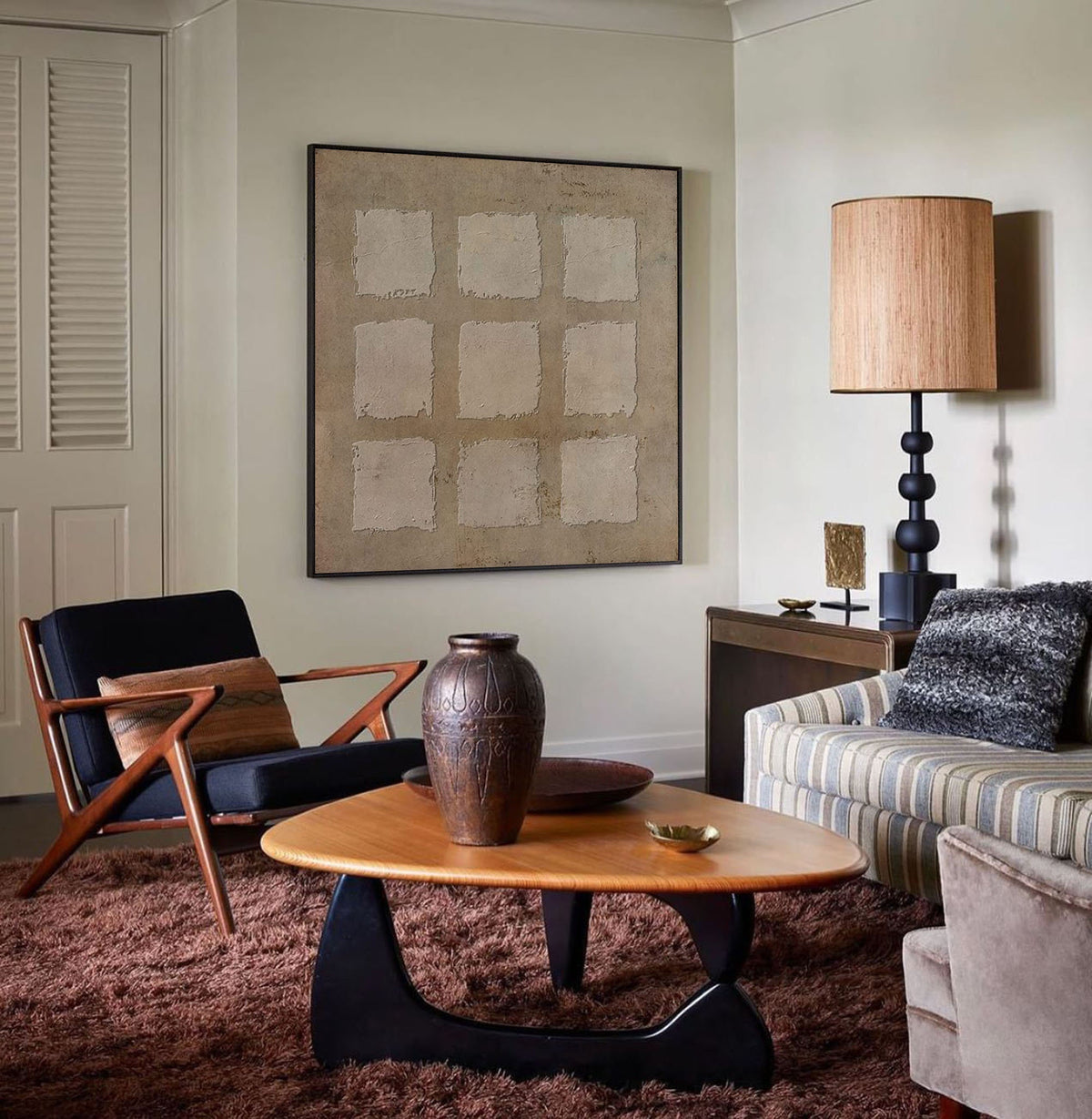 May include: A mid-century modern living room with a brown leather armchair, a coffee table with a wood top and black legs, a brown ceramic vase, a striped sofa, a lamp with a brown shade, and a framed abstract art print on the wall.