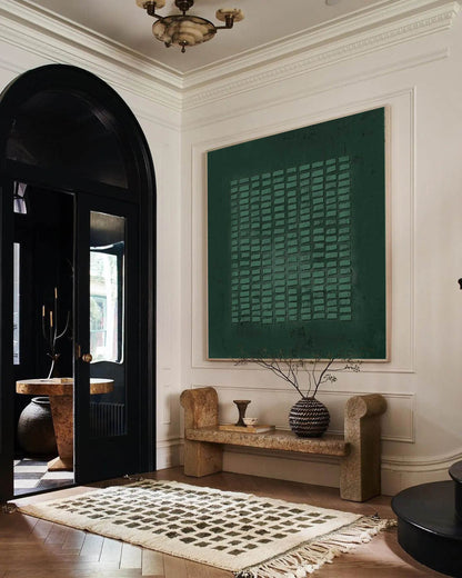 May include: A large square wall art piece featuring a dark green textured surface with a grid of raised rectangular elements. The artwork is framed in a light wood and displayed on a white wall above a stone bench with a patterned rug on the floor.