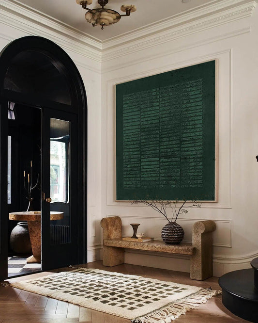 May include: A dark green abstract painting hangs above a wooden bench with a woven vase and a small decorative object. The bench is in a hallway with a black door and a patterned rug.