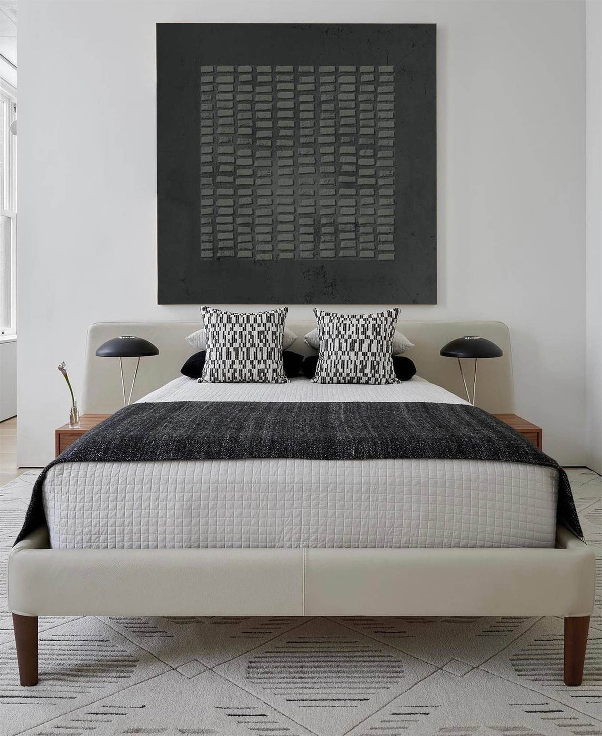 May include: A bed with a cream-colored upholstered frame and wooden legs, featuring a white quilted coverlet, a dark gray textured throw blanket, and two black and white patterned square pillows. Two black dome-shaped lamps sit on wooden nightstands.
