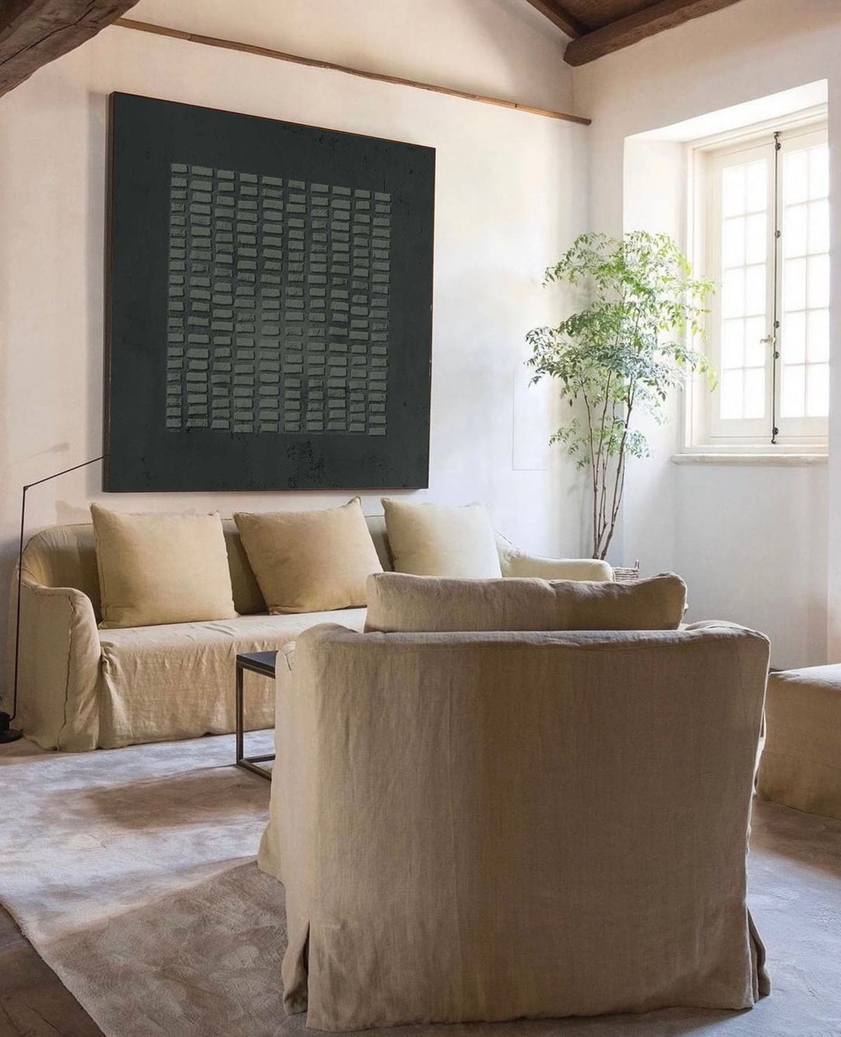 May include: A large square black wall art piece featuring a grid of small rectangular textured elements. The artwork is displayed above a beige sofa with matching cushions, positioned in a room with a beige armchair and a tall green plant.