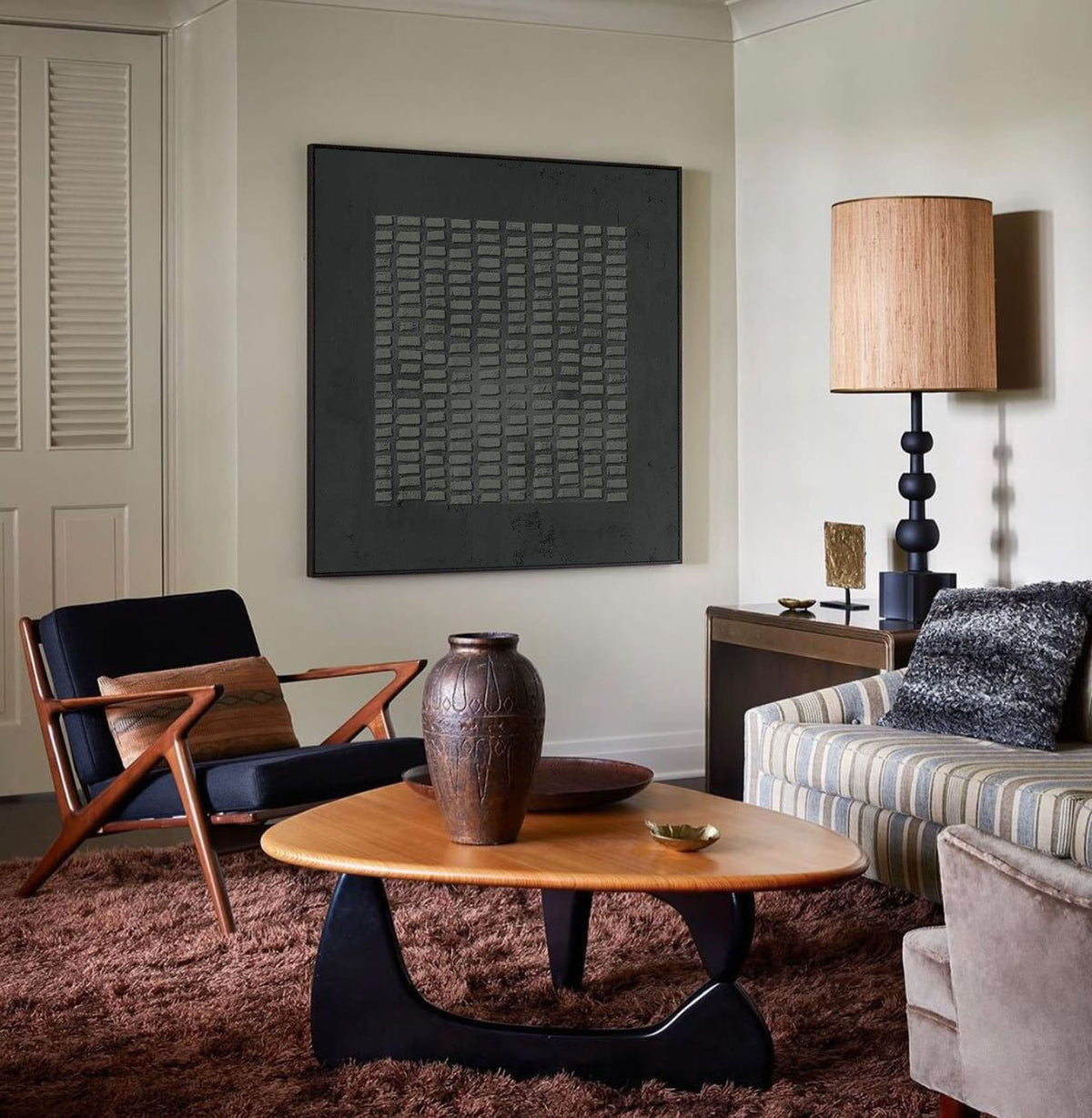 May include: A living room interior featuring a triangular wooden coffee table with a black base, a dark brown ceramic vase, a black armchair with wooden arms, a striped sofa with a textured pillow, and a large square black wall art piece.