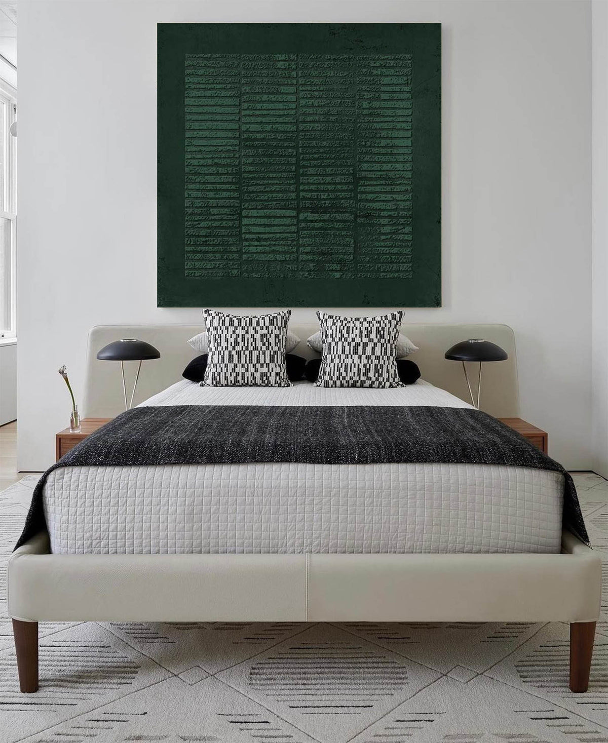May include: A white bed with a black and white patterned throw blanket and two black and white patterned pillows. A dark green abstract painting hangs above the bed.