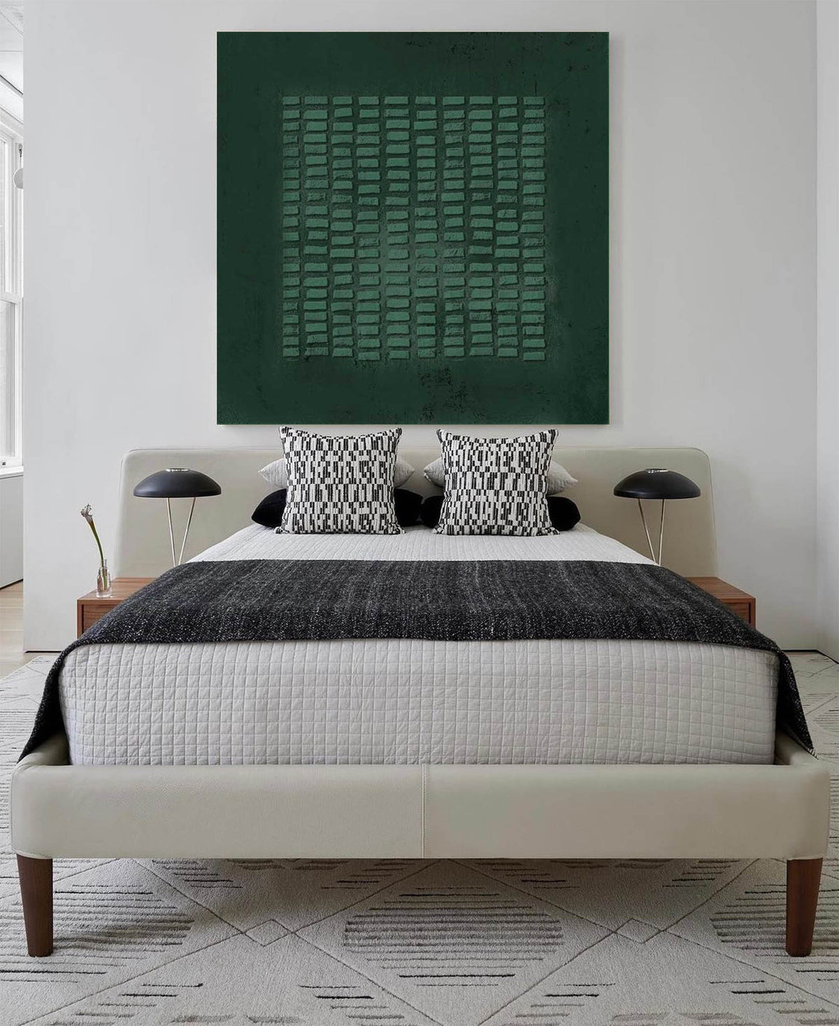 May include: A bed with a cream-colored upholstered frame and wooden legs, featuring a white quilted coverlet, a dark gray textured throw blanket, and two black and white patterned square pillows. A large dark green square wall art hangs above.