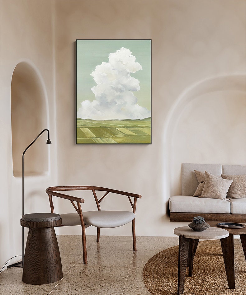 May include: A framed painting of a white cloud over a green field. The painting is hanging on a light beige wall in a room with a wooden chair and a small table.