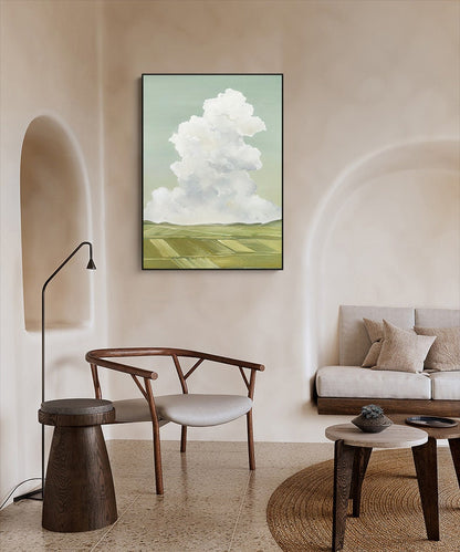 May include: A framed painting of a white cloud over a green field. The painting is hanging on a light beige wall in a room with a wooden chair and a small table.