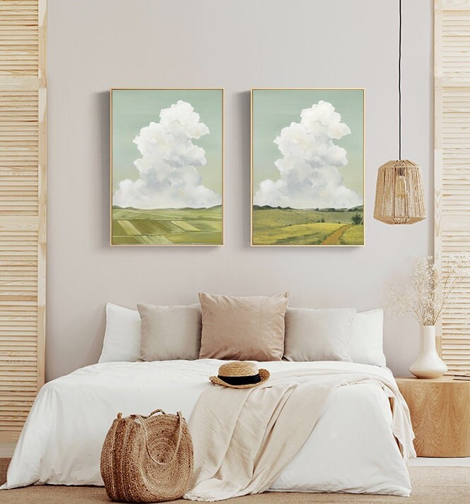 May include: Two framed landscape paintings with a green field and a white cloud in the sky. The paintings are hanging on a light gray wall above a white bed with a white blanket and a brown woven basket.