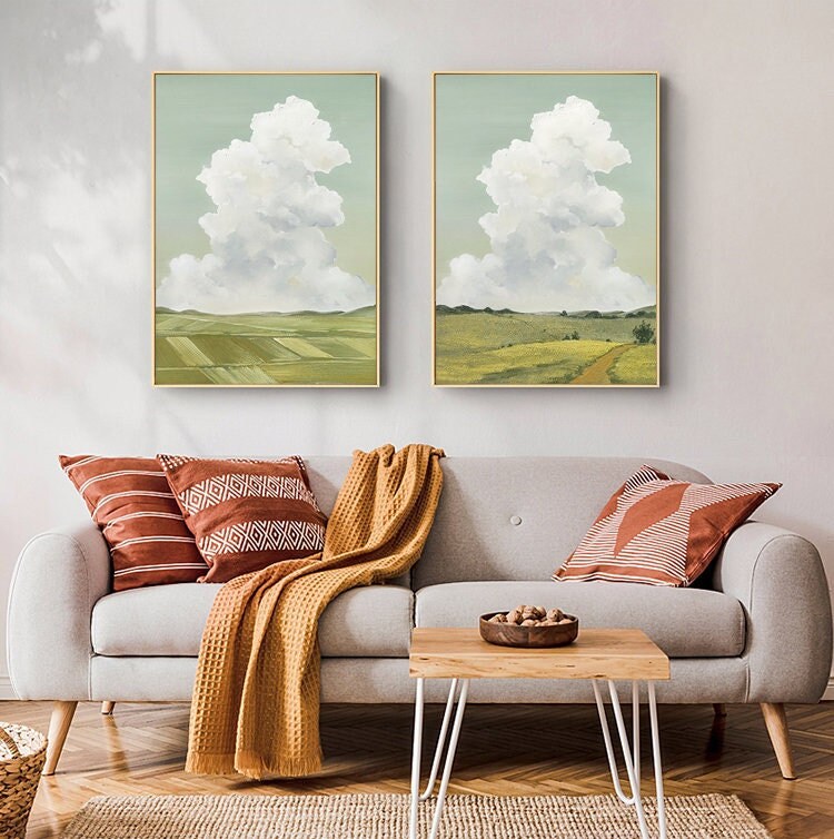 May include: Two framed landscape paintings with a green field and a large white cloud in the sky. The paintings are hanging on a white wall above a gray couch with orange throw pillows and a blanket.