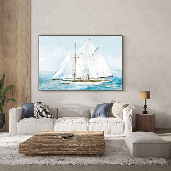 May include: A watercolor painting of a white sailboat with three sails on a blue ocean. The painting is framed and hanging on a wall above a white couch and a wooden coffee table.