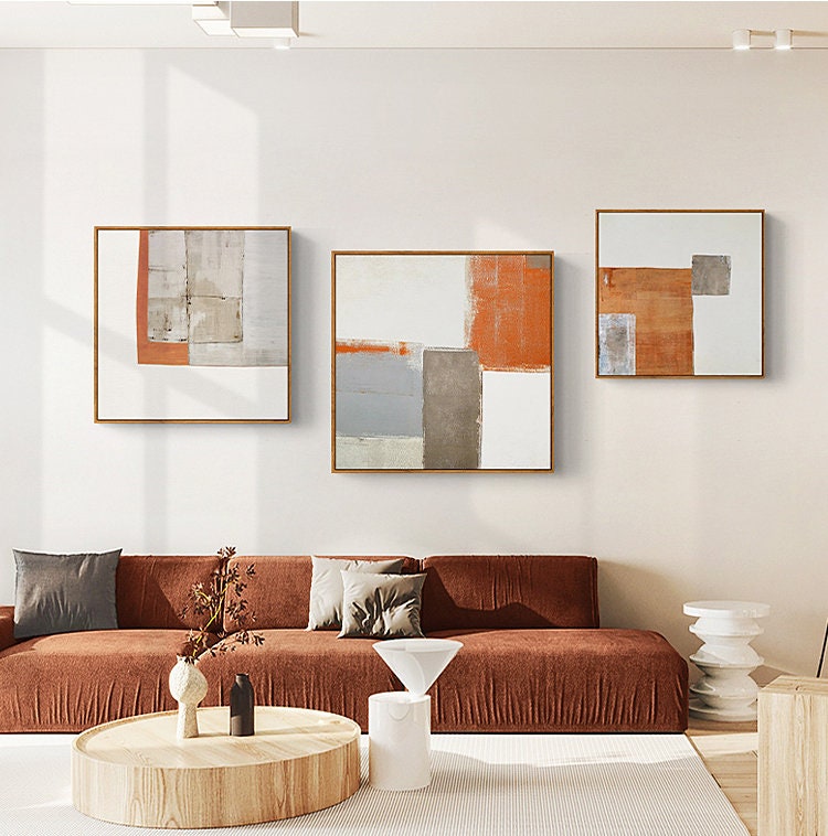 May include: Three abstract paintings with geometric shapes in shades of orange, gray, and white. The paintings are framed in a light wood frame and are hung on a white wall. The paintings are above a brown sofa with pillows.