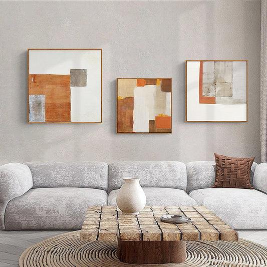 May include: Three abstract paintings in square frames with a white background and geometric shapes in shades of brown, orange, and gray. The paintings are hung on a light gray wall above a gray couch and a wooden coffee table.