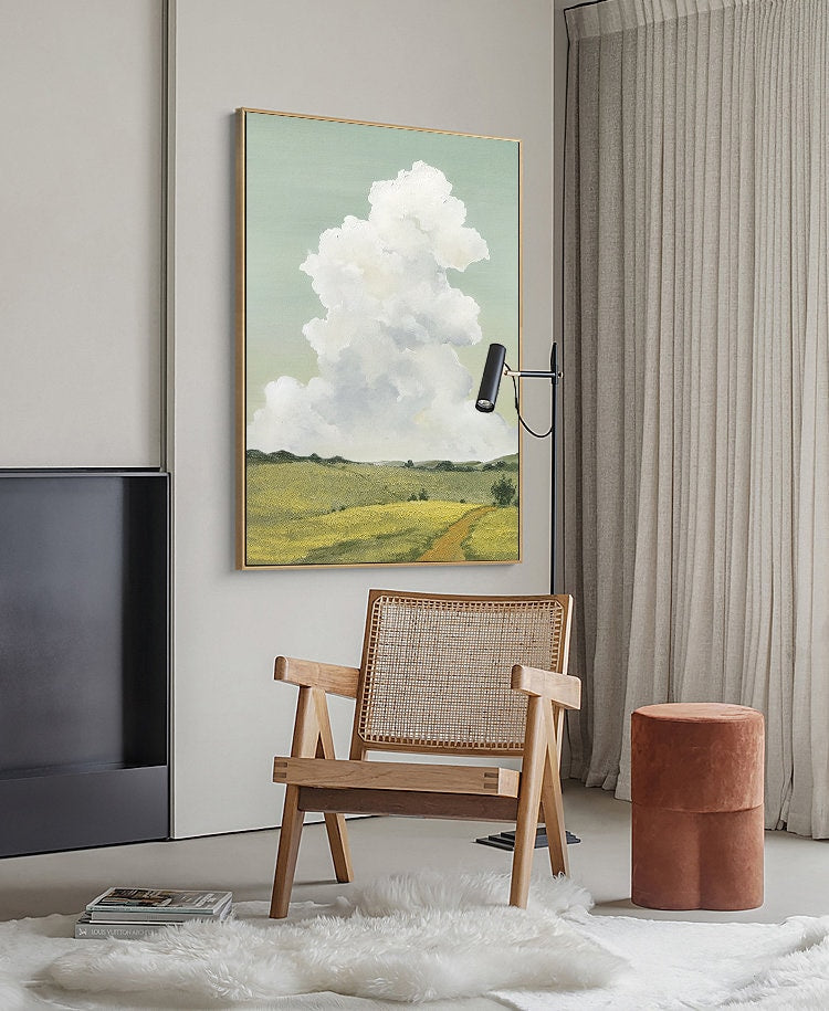 May include: A framed painting of a landscape with a large white cloud in the sky, a green field, and a dirt road leading to a small tree. The painting is hanging on a wall in a room with a wooden chair and a brown ottoman.
