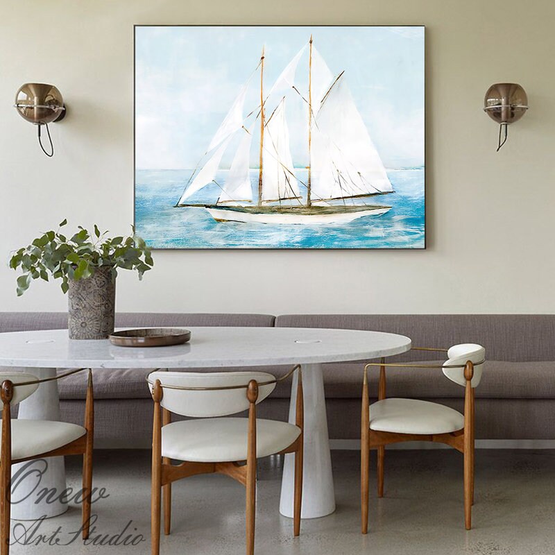 May include: A watercolor painting of a white sailboat with sails billowing in the wind, sailing on a blue ocean. The painting is framed and hanging on a wall in a room with a white marble table and chairs.