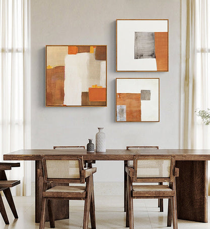 May include: Three abstract paintings with geometric shapes in shades of brown, orange, and white. The paintings are hung on a wall above a wooden dining table with chairs.