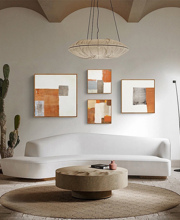 May include: Four abstract paintings with geometric shapes in shades of brown, gray, and white, hung on a white wall above a white curved sofa and a round coffee table with a textured top.