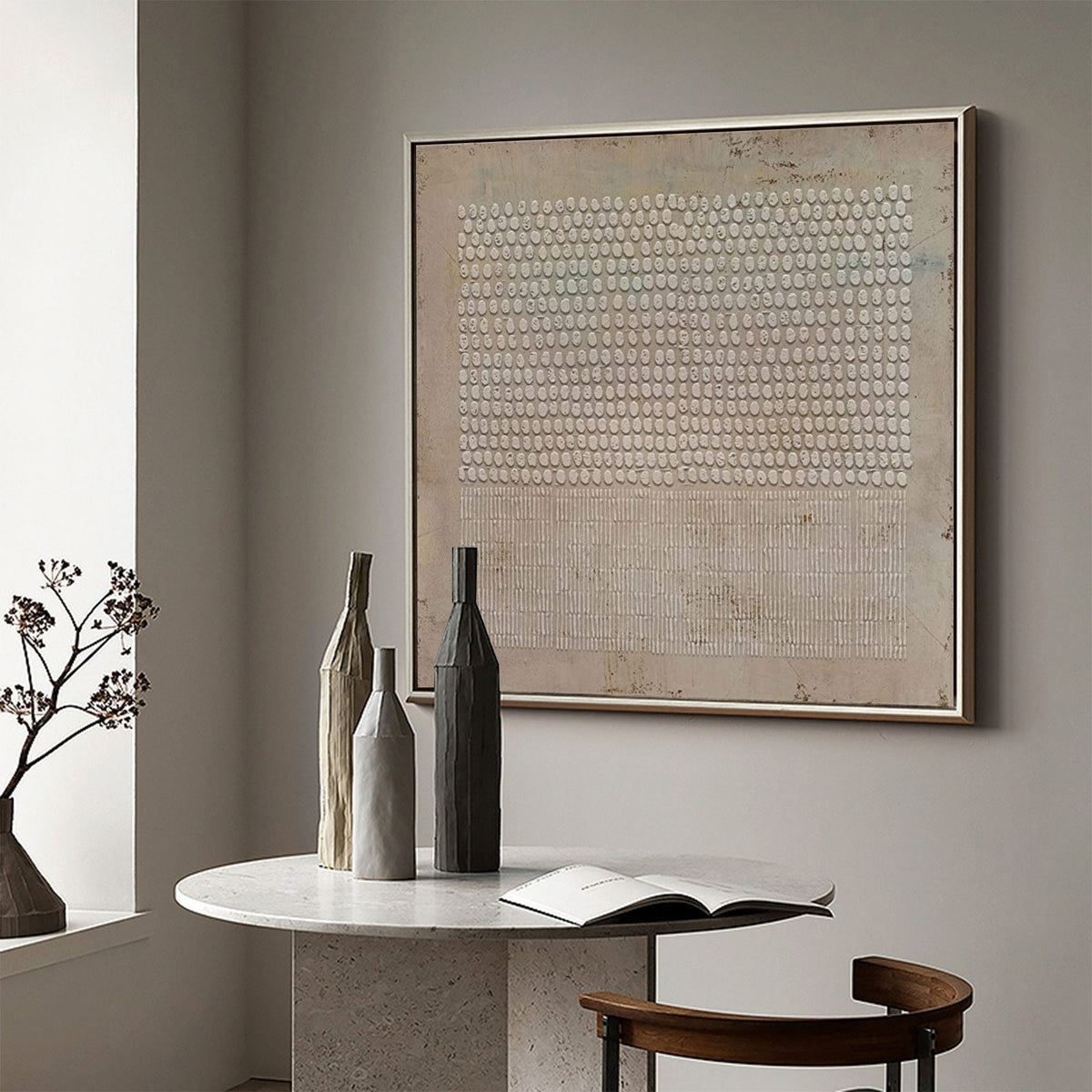 May include: Three ceramic vases in different shades of brown and beige, arranged on a white table with a book. A framed abstract painting with a pattern of white circles on a beige background hangs on the wall behind the vases.