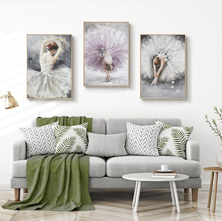 May include: Three framed paintings of ballerinas in various poses. The ballerinas are wearing white and purple tutus. The paintings are hung on a white wall above a gray couch with green throw pillows.
