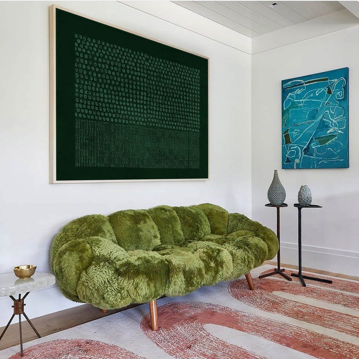 May include: A moss green, textured, fluffy sofa with a tufted design and wooden legs. Above it hangs a large, dark green rectangular artwork featuring a grid of dots and vertical lines, alongside a smaller blue abstract painting on the wall.