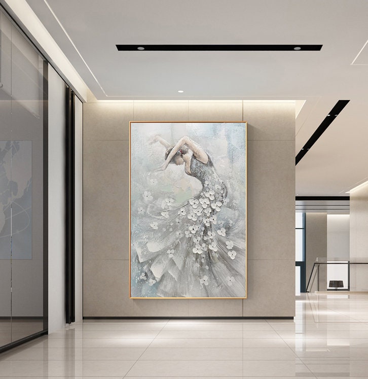 May include: An elegant painting of a dancer in a flowing gown, adorned with white floral accents. The artwork features a muted color palette of gray, white, and blue, with a gold frame. The dancer is in a graceful pose, set against a soft, textured background.