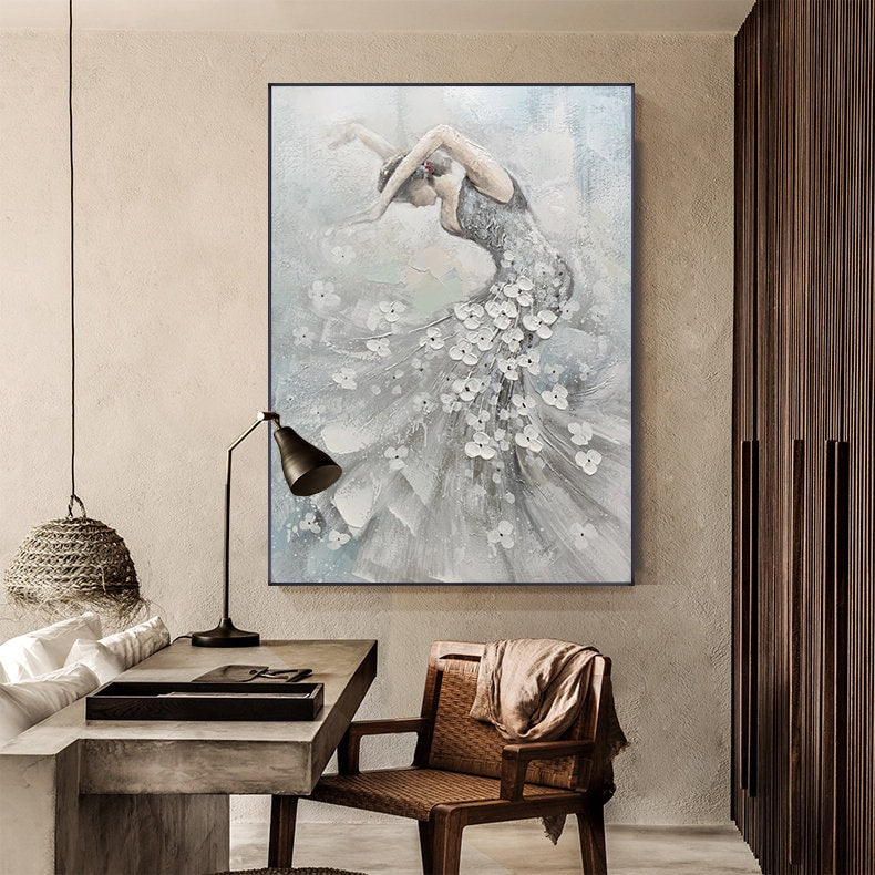 May include: An abstract painting of a dancer in a flowing gown adorned with white floral accents. The artwork features a muted color palette of gray, white, and blue, with the dancer's form gracefully arched. The painting is framed in black and hangs on a neutral-toned wall.
