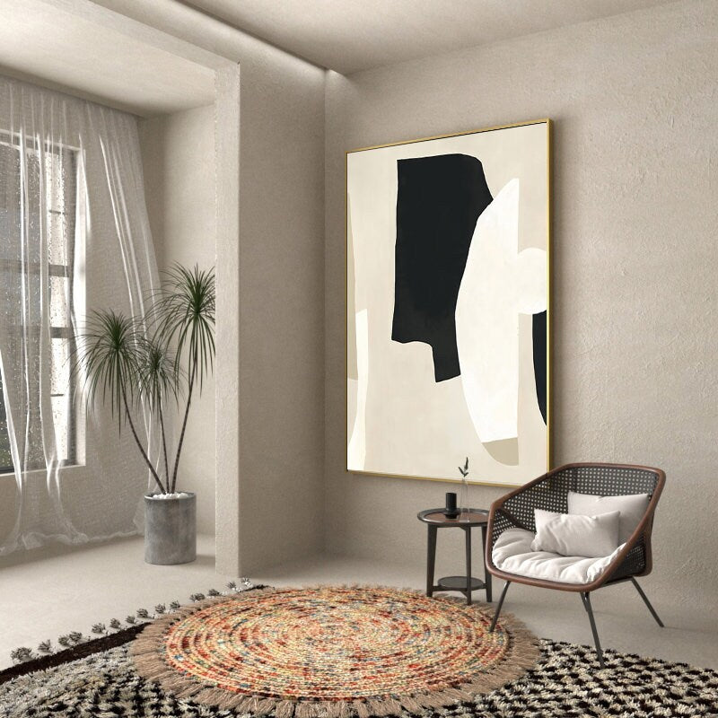 May include: A modern abstract painting with black and white shapes on a beige background. The painting is framed in gold and hangs on a light beige wall. A round rug with a fringe border is on the floor in front of a chair with a white cushion.