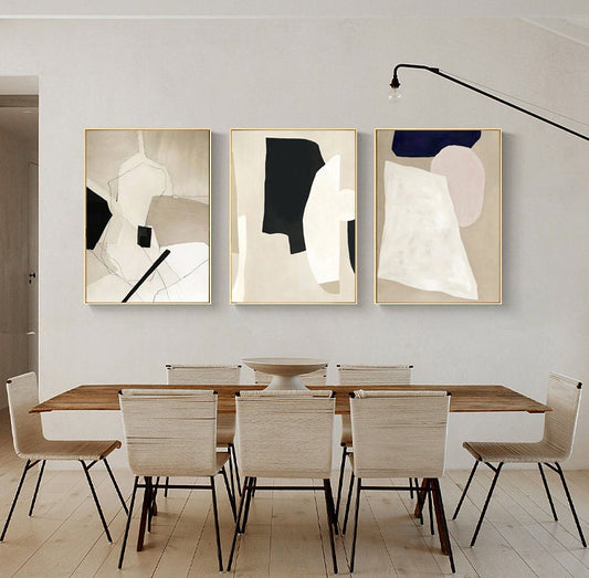 May include: Three abstract paintings in gold frames with black, white, and beige geometric shapes. The paintings are hanging on a white wall above a wooden dining table with six chairs.