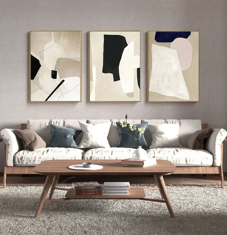 May include: Three abstract art prints with geometric shapes in black, white, beige, and pink. The prints are framed in gold and hung on a light gray wall above a white sofa with pillows.