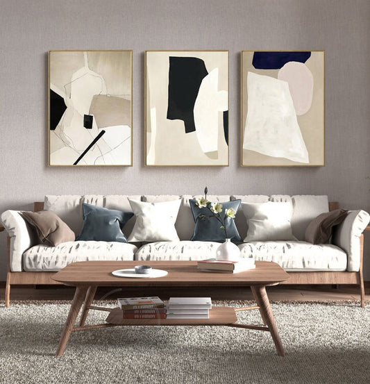 May include: Three abstract art prints with geometric shapes in black, white, beige, and pink. The prints are framed in gold and hung on a light gray wall above a white sofa with pillows.