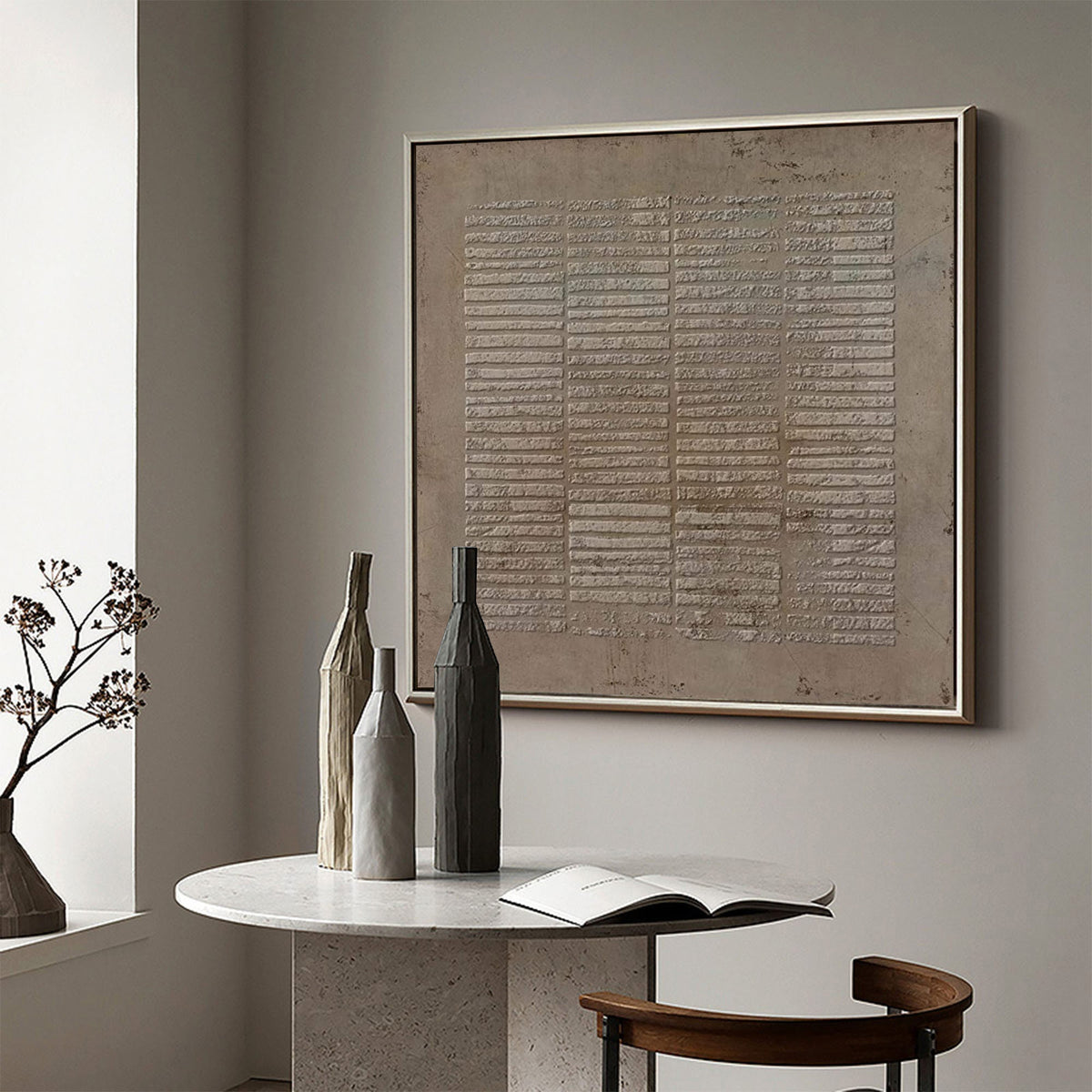 May include: A square framed wall art piece featuring a textured, beige, grid-like pattern of horizontal lines. Below, a round white marble table holds three tall, slender, cylindrical vases in beige, light gray, and dark charcoal gray colors.