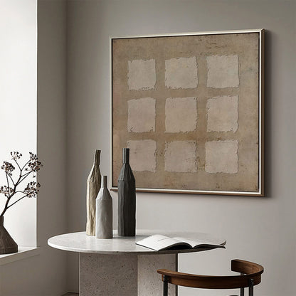 May include: Three ceramic vases in different shapes and colors, arranged on a white table in front of a beige wall with a framed abstract painting. The painting features a grid of nine squares with a textured, beige background.