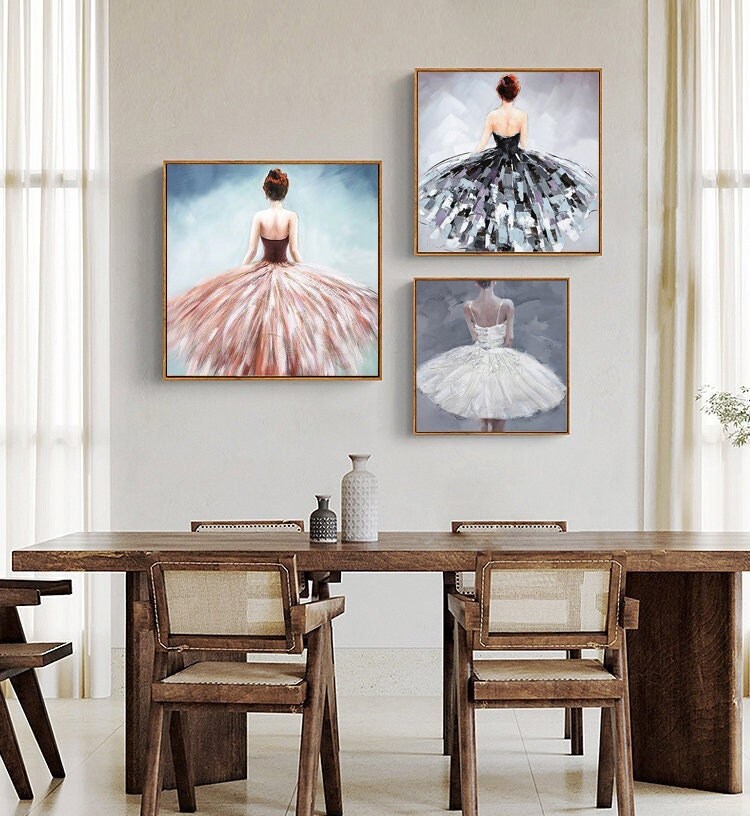 May include: Three framed paintings of ballerinas wearing different colored tutus. The ballerinas are facing away from the viewer and are all wearing their hair up. The paintings are hanging on a light gray wall above a wooden table with chairs.