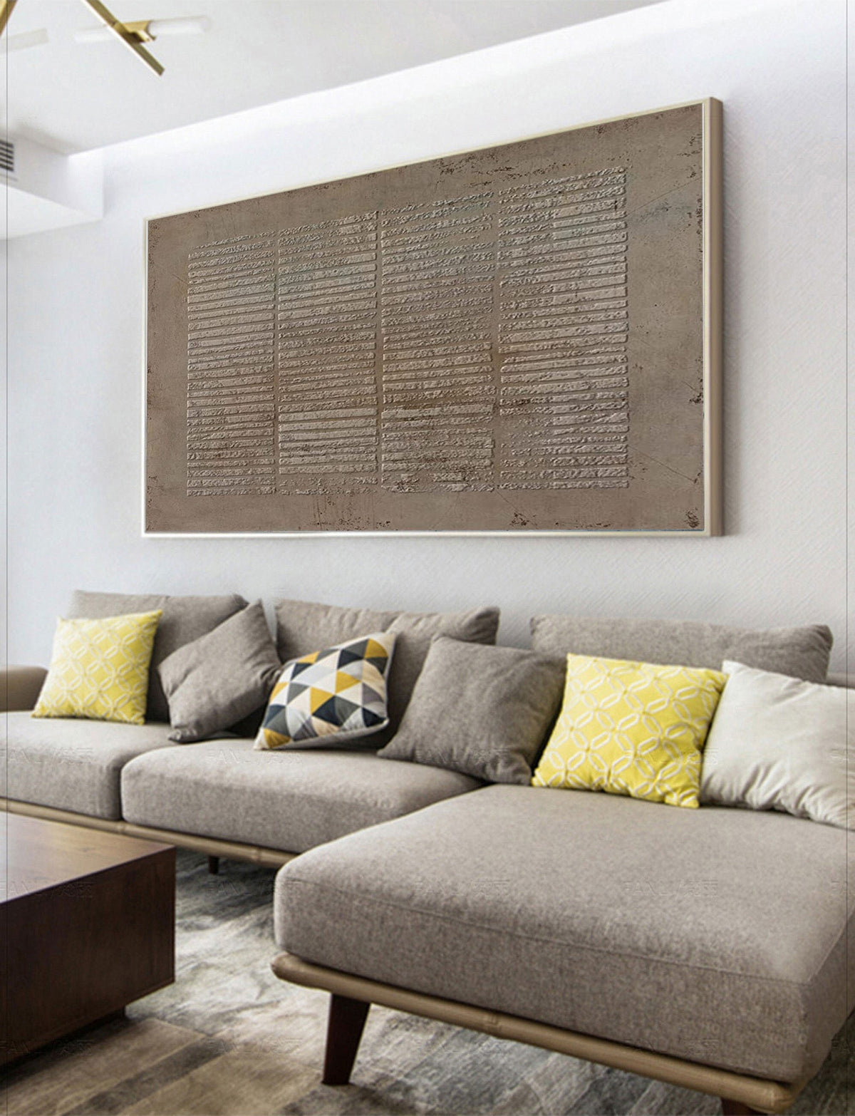 May include: A large, rectangular, abstract painting with a textured, gray surface. The painting is framed in a thin, silver frame and is hanging on a white wall above a gray sectional sofa with yellow and gray throw pillows.