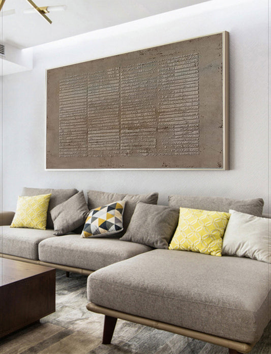 May include: A large, rectangular, abstract painting with a textured, gray surface. The painting is framed in a thin, silver frame and is hanging on a white wall above a gray sectional sofa with yellow and gray throw pillows.