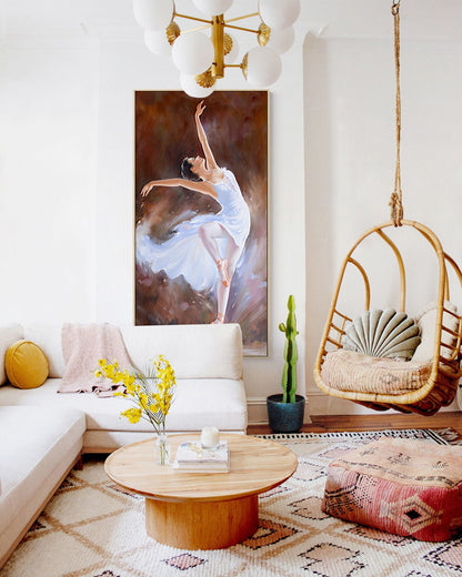 May include: A white couch with a yellow throw pillow sits in a living room with a white rug. A wooden coffee table with a vase of yellow flowers and a book sits in front of the couch. A hanging wicker chair with a light green pillow hangs from the ceiling. A painting of a ballerina in a white dress hangs on the wall above the couch. A cactus plant sits in a blue pot on the floor.