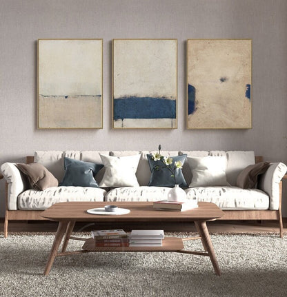 May include: Three abstract paintings in gold frames with a white background and blue accents hang above a white sofa with brown and blue pillows. A wooden coffee table with a lower shelf sits in front of the sofa.