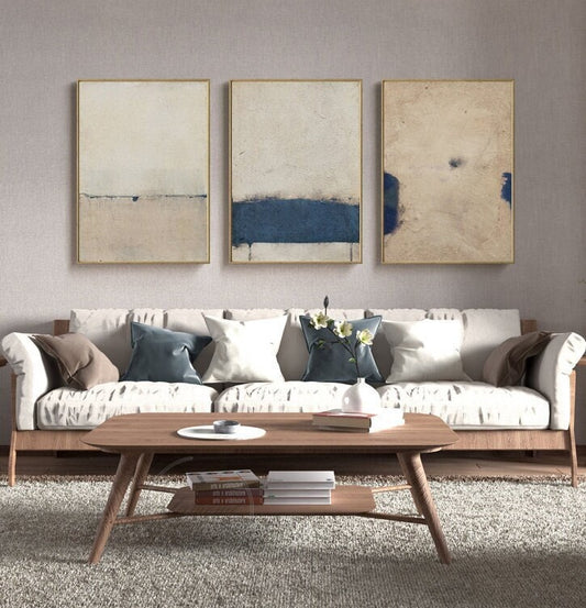 May include: Three abstract paintings in gold frames with a white background and blue accents hang above a white sofa with brown and blue pillows. A wooden coffee table with a lower shelf sits in front of the sofa.