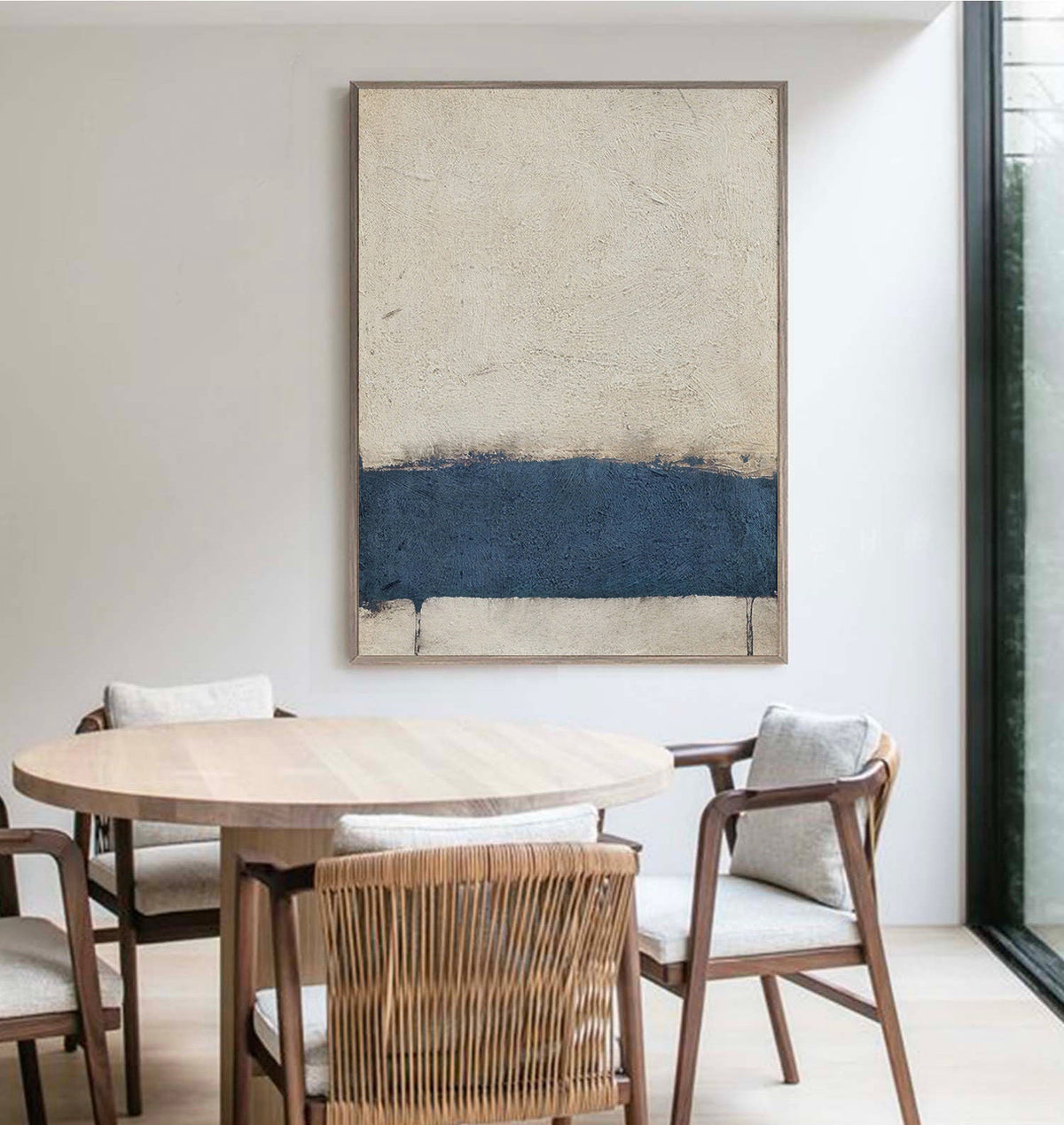 May include: Abstract painting with a horizontal band of dark blue paint on a light beige background. The painting is framed and hanging on a white wall above a wooden table with chairs.