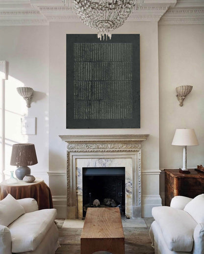 May include: A large, dark gray abstract artwork hangs above a fireplace in a living room. The artwork features a textured, vertical stripe pattern. The room has white walls, a chandelier, and classic furniture.