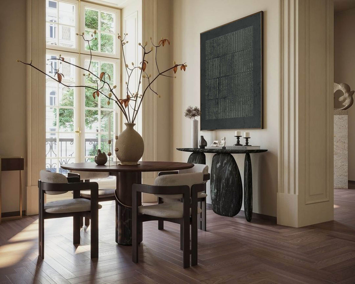 May include: A dining room scene featuring a round wooden table with four chairs. A large vase with branches and leaves sits in the center. A dark abstract painting hangs on the wall, and a console table with a stone base is also present.