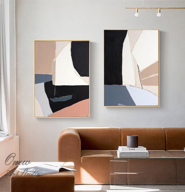 May include: Two abstract paintings with geometric shapes in shades of brown, white, black, and blue. The paintings are framed in gold and hung on a white wall.