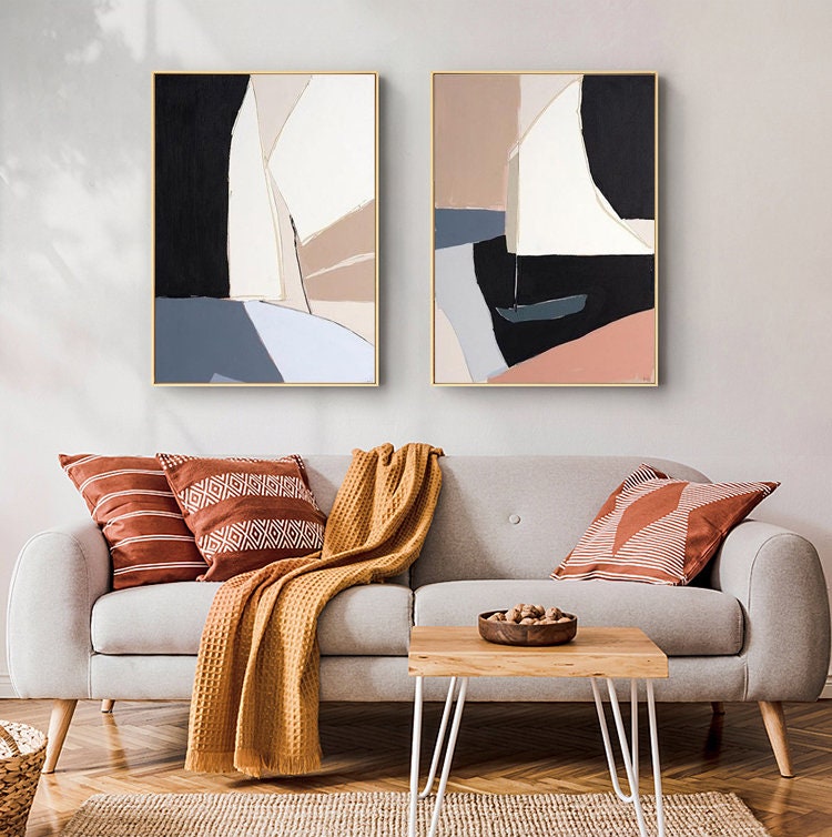 May include: Two abstract paintings with geometric shapes in shades of black, white, beige, pink, and blue. The paintings are framed in gold and hung on a white wall above a gray sofa with throw pillows and a blanket.
