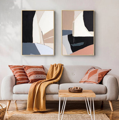 May include: Two abstract paintings with geometric shapes in shades of black, white, beige, pink, and blue. The paintings are framed in gold and hung on a white wall above a gray sofa with throw pillows and a blanket.