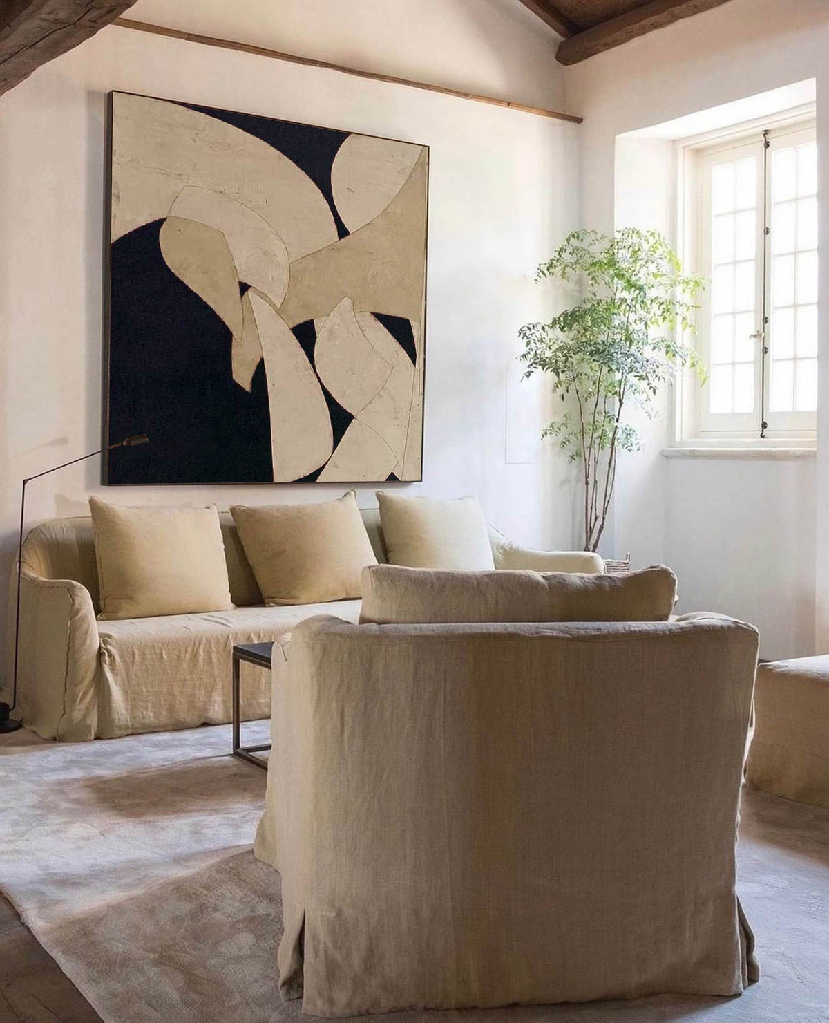May include: A living room scene featuring a large abstract painting with black, beige, and white shapes. A beige sofa with matching pillows and slipcovers is in front of the painting. Two beige armchairs and a small table complete the room.