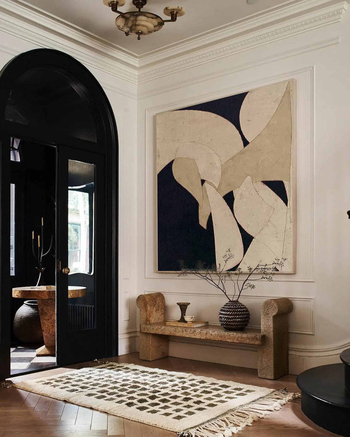 May include: An interior shot of a hallway with a large abstract painting in neutral tones. A stone bench with a decorative vase and a patterned rug add to the room's aesthetic. A black arched doorway leads to another room.