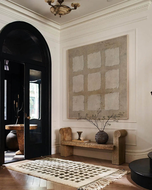 May include: A white rug with a geometric pattern is placed in front of a black door with a large arched window. A wooden bench with a woven basket and a small vase sits in front of a large abstract painting with a beige background.