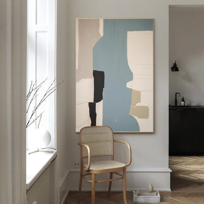 May include: Abstract art painting with geometric shapes in shades of beige, black, white, and blue. The painting is hanging on a white wall in a room with a wooden chair and a window.